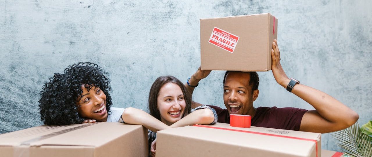 Two smiling women and a man surrounded by cardboard boxes. The man holds a box over his head.